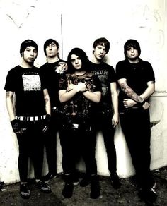 four young men standing next to each other in front of a white wall with graffiti on it