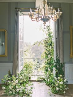 an open window with flowers and greenery in front of it, next to a chandelier