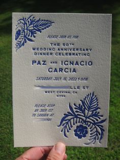 a hand holding up a wedding card in front of the camera on some green grass