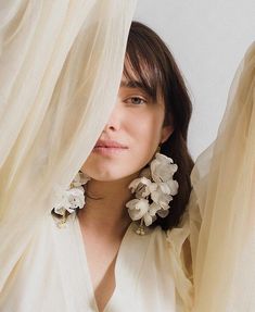 a woman with flowers on her ear stands in front of sheer curtains