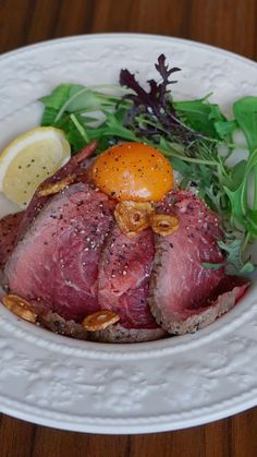 a white plate topped with meat and vegetables on top of a wooden table next to a slice of lemon