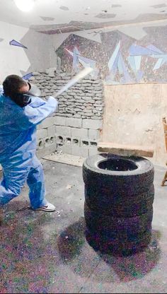 a man in blue coveralls swinging a baseball bat at a black barrel with graffiti on the wall behind him