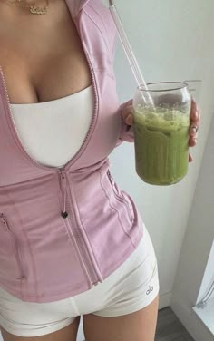 a woman is holding a green smoothie in her right hand and wearing white shorts