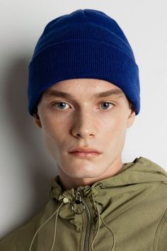 a young man wearing a blue beanie and green jacket with his head tilted to the side