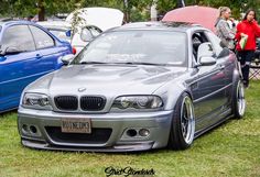 a silver car parked next to other cars on top of a grass covered field in front of people