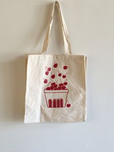 "This 100% cotton tote bag features a hand drawn basket full of cherries. This bag is good for the farmer's market, as a grocery tote bag, for school or every day use. It is printed here in dark red ink. We screen print these tote bags to order in our shop in Connecticut. Since these tote bags are each hand crafted and made to order, there may be slight variations in placement, but they are all beautiful! // FABRIC: 100% cotton // COLOR: Printed with red ink. Bag color is natural cotton (off-whi Reusable Cotton Bags For Grocery Shopping, Reusable Cotton Grocery Shopping Bags, Organic Cotton Canvas Bag For Gift, Eco-friendly Red Cotton Canvas Bag, Eco-friendly Cotton Bags For Farmers Market, Natural Cotton Grocery Bag, Natural Cotton Grocery Bags, Organic Cotton Bags For Gift, Organic Cotton Bag For Gift