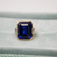 This exquisite 12 Ct Lab Grown Emerald Cut Sapphire and Trapezoid Diamond Ring is masterfully set in 18K yellow gold. This piece captures the essence of modern elegance with its sharp, clean lines and a striking bezel setting. Flanked by trapezoid diamonds, the sapphire centerpiece exudes a royal blue hue. Perfect for those who cherish contemporary design with a hint of classic allure. Three Stone Ring, Three Stone Rings, Modern Elegance, Three Stone, Blue Hues, Stone Ring, Bezel Setting, Emerald Cut, Clean Lines