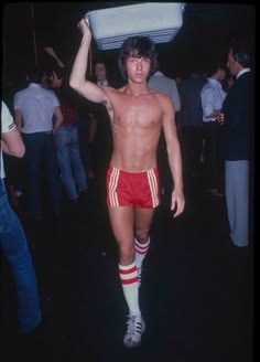 a shirtless man carrying a cooler over his head at a party with other people in the background