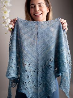 a woman holding up a blue crocheted shawl with flowers in the background