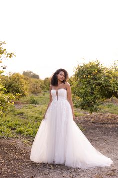 a woman in a white wedding dress standing on a dirt road with trees and bushes behind her