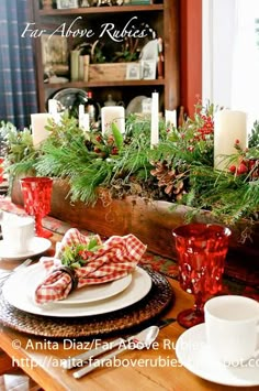 the table is set for christmas dinner with red and white dishes, candles and greenery