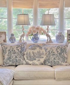 a white couch with blue and white pillows on it in front of two windows that have vases filled with flowers