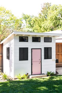 a white shed with pink door and black shutters on the side, text overlay reads how to build a modern wooden playhouse