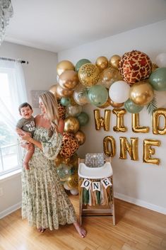 a woman holding a child in front of a balloon wall with wild one on it