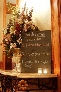 a welcome sign on a table with flowers and candles in front of the chalkboard