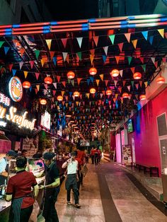 people are walking through an open market with colorful lights hanging from the ceiling above them