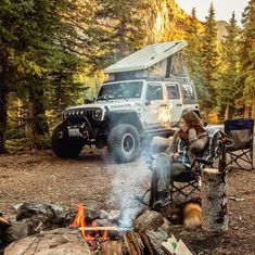 a woman sitting in a chair next to a campfire with a jeep parked behind her