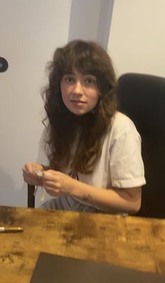 a woman sitting in front of a wooden table holding a remote control and looking at the camera