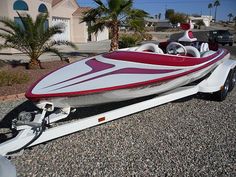 a red and white speed boat is parked on the gravel next to a palm tree