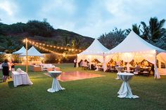 a group of people standing around tables and chairs under white tents with lights on them