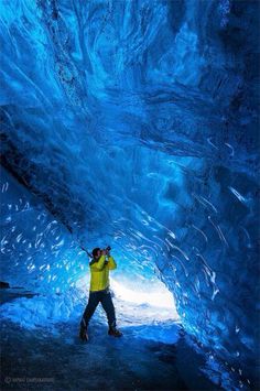 a man in yellow jacket standing inside of a blue ice cave with water flowing down it