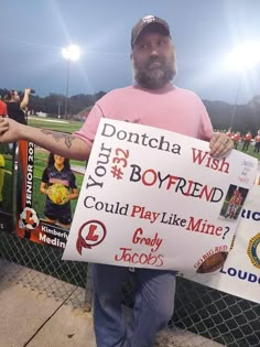 a man holding up a sign in front of a fence at a baseball field that says, dontcha wish boyfriend could play like mine