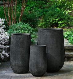 three large black vases sitting on top of a stone floor next to trees and bushes