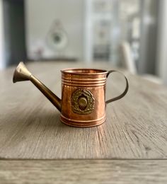 a metal watering can sitting on top of a wooden table next to a cup with a handle