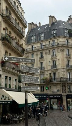 street signs pointing in different directions on a cobblestone road with buildings behind them