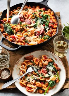 a pan filled with pasta and spinach on top of a wooden table next to two glasses