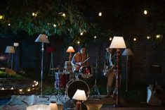 two men playing instruments in the dark with lights around them and lamps on either side