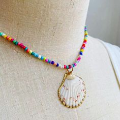 Embrace the essence of summer with this exuberant, colorful beaded necklace, featuring a stunning natural seashell pendant encased in a delicate golden trim. Each bead is meticulously selected to create a kaleidoscope of joy around your neck, reminiscent of a sun-kissed day at the beach or a vibrant festival dance. The heart of this necklace is the seashell pendant, a treasure from the ocean's depths, polished to perfection, and framed in gold. This piece is a celebration of nature's artistry, w Shell-shaped Beaded Necklaces For Beach Season, Adjustable Shell Beaded Necklace With Colorful Beads, Bohemian Shell With Colorful Beads, Ocean-inspired Beaded Necklace For Summer, Summer Ocean-inspired Beaded Necklaces, Beach Shell Necklaces With Tiny Beads, Beach Shell Necklaces With Colorful Beads, Beaded Shell-shaped Jewelry For The Beach, Colorful Beaded Shell Necklaces For Vacation