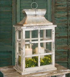 a white lantern with two lit candles inside on a wooden table in front of a green wall