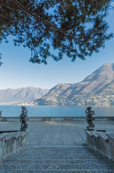 a stone walkway with statues on each side and mountains in the background, along with blue water