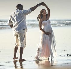 a pregnant woman in a white dress dancing on the beach next to a man with his arm around her waist