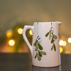 a white pitcher with green leaves on it sitting on a table next to a christmas tree