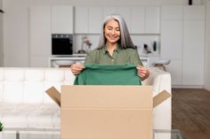 an older woman holding a green shirt in front of a cardboard box on a couch