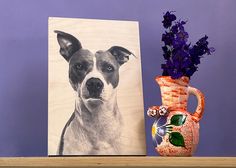 a painting of a dog next to a vase with purple flowers in it and a photo on the wall