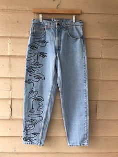 a pair of blue jeans with hand drawn faces on them hanging from a wooden hanger