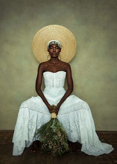 a woman in a white dress and straw hat sitting on the floor with plants around her