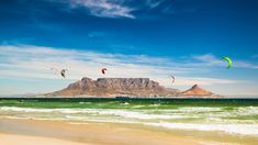 several kites are flying over the ocean on a sunny day with mountains in the background