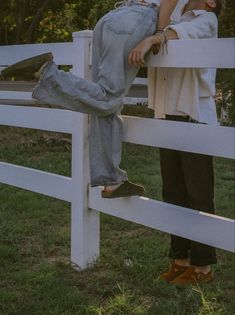 two people sitting on top of a white fence next to each other in the grass