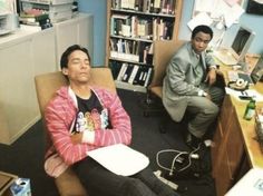 two men sitting in an office with computers on the desk and bookshelves behind them