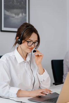 a woman sitting at a desk with a laptop and headphones on her ears, talking on the phone