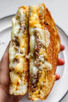 a hand holding a sandwich with cheese and meat on it in front of a white plate