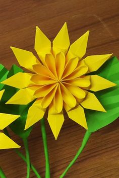 two yellow paper flowers sitting on top of a wooden table next to green stems and leaves