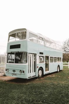 a double decker bus parked on the grass in front of a white fence and trees