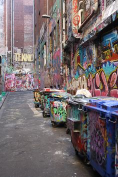 graffiti painted on the side of buildings in an alleyway with garbage cans lined up next to each other