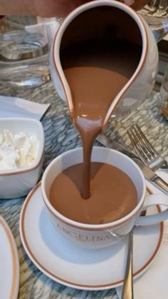 chocolate being poured into a cup on a table with other plates and silver utensils