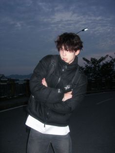 a man with his arms crossed standing in the street at night, wearing a black jacket and white t - shirt